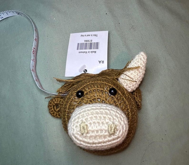 May include: A brown and cream-colored crochet tape measure shaped like a horse's head. The tape measure is white with black markings. A tag attached to the tape measure reads "This is not a toy" and "Made in Vietnam".