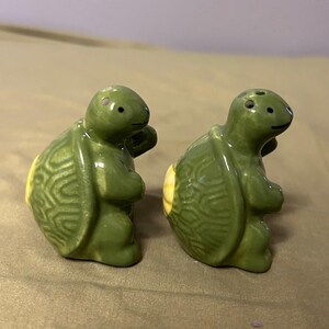 Puede incluir: Un par de saleros y pimenteros con forma de tortuga de cerámica. Las tortugas son verdes con detalles amarillos en sus caparazones. El salero tiene agujeros en la parte superior. Los caparazones de las tortugas tienen un diseño texturizado.