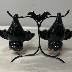 May include: A pair of glossy black ceramic bat-shaped salt and pepper shakers hanging upside down from a black metal stand. The stand features a small decorative bat at the top center. Each bat shaker has three small holes on its back.