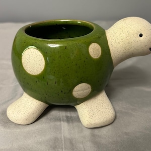 Puede incluir: Una maceta de cerámica con forma de tortuga. El cuerpo es verde con lunares blancos, y la cabeza y las patas son blancas con detalles moteados. La maceta tiene una abertura redonda para plantas y una cara sonriente.