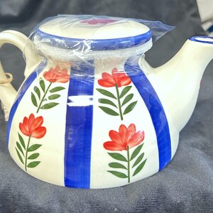 May include: A cream-colored ceramic teapot featuring vertical cobalt blue stripes and a repeating pattern of red flowers with green stems and leaves. The teapot has a matching lid, a curved handle, and a spout, with a thin twine tag attached.