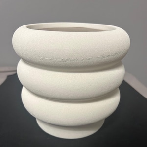 Puede incluir: Un jarrón de cerámica blanco texturizado con un diseño apilado y redondeado. El jarrón tiene una abertura ancha y un color neutro, adecuado para varios estilos de decoración. El jarrón mide unos 20 cm de alto.