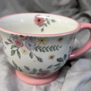 May include: A white ceramic teacup with a pink handle and base. The cup is decorated with a floral pattern in pink, blue, and yellow. The rim of the cup is also pink.