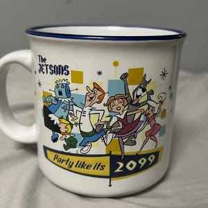 May include: White ceramic mug featuring a colorful illustration of The Jetsons cartoon characters. The mug has a blue rim and a handle. Text on the mug reads "The Jetsons" and "Party like its 2099".