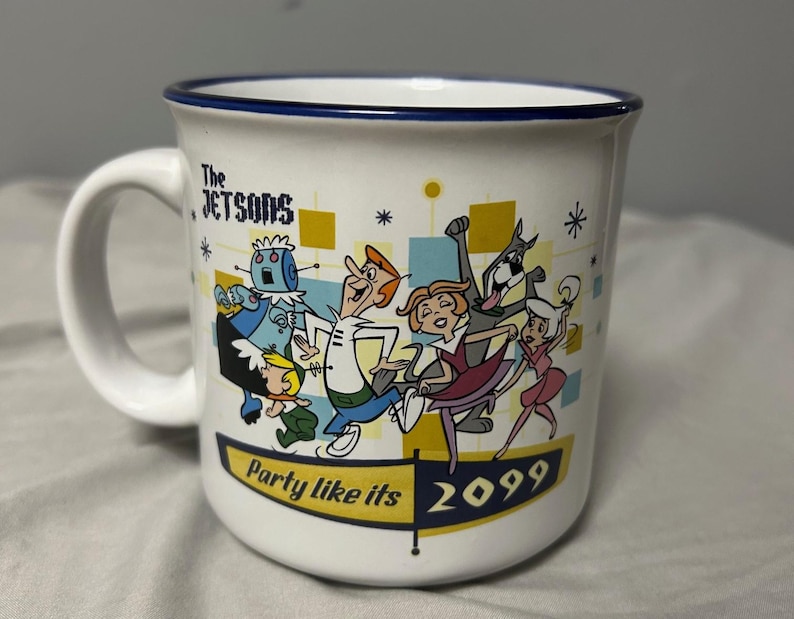 May include: White ceramic mug featuring a colorful illustration of The Jetsons cartoon characters. The mug has a blue rim and a handle. Text on the mug reads "The Jetsons" and "Party like its 2099".