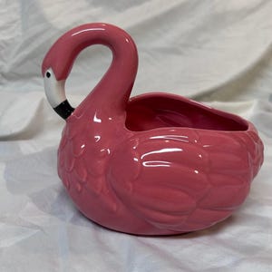 May include: A pink ceramic flamingo planter with a curved neck and open basin. The flamingo has a white beak with a black tip and a detailed feather design. The planter is a decorative item.