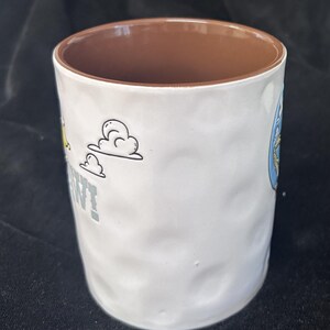 Pu&ograve; includere: Tazza in ceramica bianca con esterno strutturato e interno marrone. La tazza presenta grafiche di cartoni animati e la parola "WANT" in lettere grigie. La tazza &egrave; cilindrica e misura circa 10 cm di altezza.