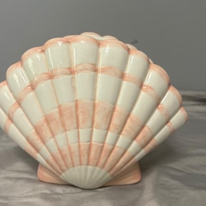 May include: A ceramic seashell sculpture in a fan shape. The shell is primarily white with alternating bands of light pink. The sculpture has a smooth, glossy finish and is designed to stand upright.