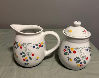 May include: A white ceramic creamer and sugar set with a floral design. The creamer has a handle and a spout. The sugar bowl has a lid. Both pieces feature a pattern of yellow, red, blue, and green flowers on a white background.