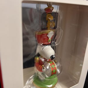 May include: A Snoopy and Woodstock Christmas ornament inside a white box. Snoopy wears a red and gold soldier uniform with a tall hat and plays a drum with green and red diamond patterns. Woodstock sits on top of Snoopy's hat wearing a matching hat.