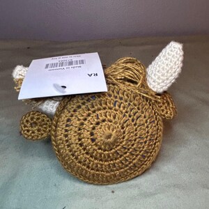 May include: A crocheted, round, brown object with white ears and a small, round, brown ear on the side. A tag is attached to the top. The object is made of yarn and has a spiral design.