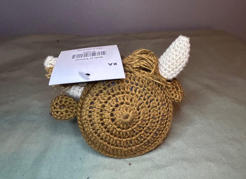 May include: A crocheted, round, brown object with white ears and a small, round, brown ear on the side. A tag is attached to the top. The object is made of yarn and has a spiral design.