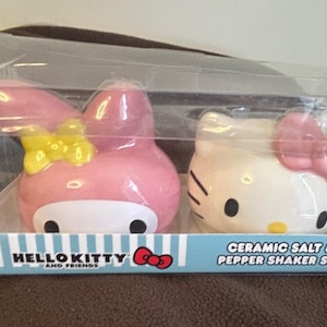 Puede incluir: Un juego de salero y pimentero de cerámica de Hello Kitty and Friends. El juego incluye dos saleros con forma de personaje, uno rosa con un lazo amarillo y otro blanco con un lazo rosa. La caja dice "CERAMIC SALT & PEPPER SHAKER SET."