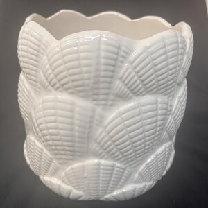 May include: A white ceramic planter featuring a repeating embossed shell pattern across its exterior surface. The vessel has a scalloped top rim and a cylindrical shape, with a glossy finish that highlights the textured details of the shells.