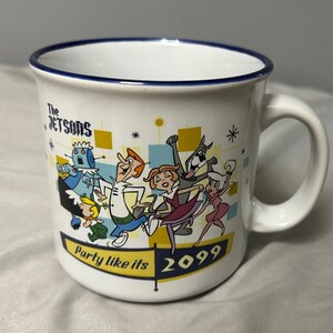 May include: White ceramic mug featuring The Jetsons cartoon characters. The mug has a blue rim and a handle. The design includes the text "Party like its 2099" and cartoon characters in a retro style.