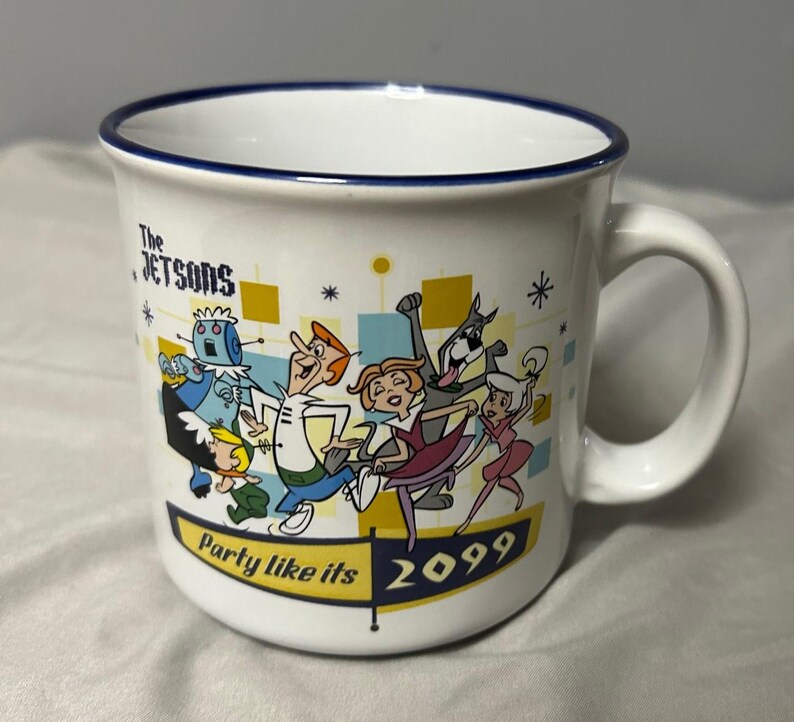 May include: White ceramic mug featuring The Jetsons cartoon characters. The mug has a blue rim and a handle. The design includes the text "Party like its 2099" and cartoon characters in a retro style.