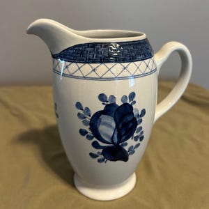 Puede incluir: Jarra de cerámica blanca con diseño floral azul. La jarra presenta una banda azul tejida alrededor de la parte superior y un motivo floral azul en el cuerpo. Tiene un asa curva y un pico para verter.