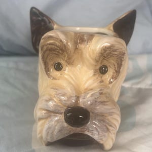 May include: A ceramic mug shaped like the head of a terrier dog. The mug features a cream-colored face with brown painted details around the eyes, ears, and muzzle. It has a glossy finish, a black nose, and two upright ears with dark brown tips.