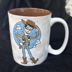 Pu&ograve; includere: Tazza in ceramica bianca con interno marrone e manico grande. La tazza presenta una colorata illustrazione di Woody di Toy Story, su uno sfondo blu con nuvole. Un oggetto da collezione divertente.