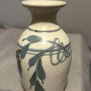 Op de afbeelding: Een kleine, crèmekleurige keramische vaas met een uitlopende rand en een afgeronde body. De vaas is versierd met een blauw blad- en lintontwerp. De vaas is ongeveer 15 cm hoog.