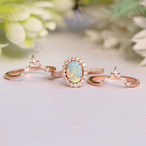 May include: A three-piece rose gold ring set. The central ring has an oval opal surrounded by a halo of small diamonds. Two additional bands with diamond accents complete the set. The rings are displayed on a white surface.