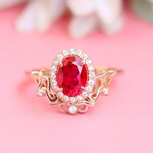 May include: A rose gold engagement ring with a large oval ruby surrounded by a halo of smaller diamonds. The ring band features a delicate, intricate design.