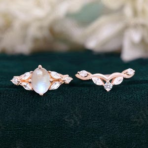 Puede incluir: Un conjunto de anillo de compromiso de oro rosa con una piedra central de piedra de luna de forma de pera y una banda de boda de oro rosa a juego con un diseño curvo y pequeños diamantes.