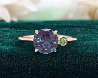 May include: A rose gold ring with a large, round, blue gemstone and a smaller, round, green gemstone. The ring is set on a green velvet background.