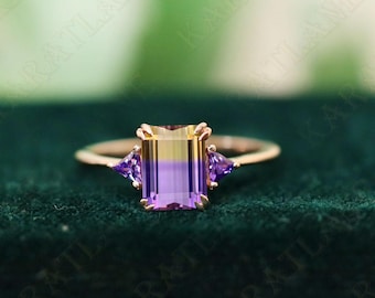 May include: A gold ring with a large, emerald-cut gemstone. The gemstone is a gradient of yellow and purple. The ring also has two smaller, triangular gemstones on either side of the main stone.
