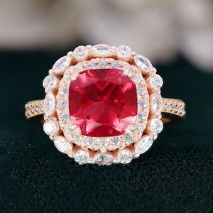 May include: A rose gold ring with a large, square-cut red gemstone surrounded by a double halo of smaller, round white gemstones.
