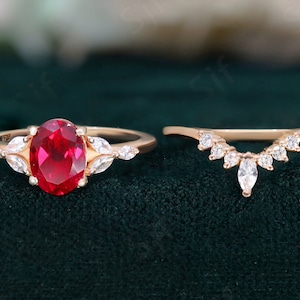 May include: A rose gold engagement ring set with a large oval ruby surrounded by smaller diamonds. The matching wedding band is a delicate rose gold band with a row of small diamonds.
