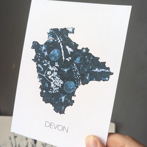 May include: A white card with a blue outline of the county of Devon, England. The outline is filled with a blue and white floral pattern. The word "DEVON" is printed in black at the bottom of the card.