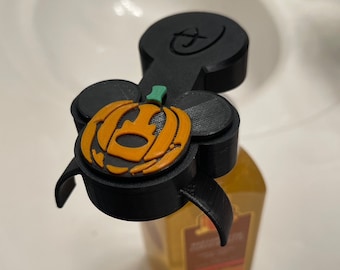 May include: Black plastic bottle stopper shaped like Mickey Mouse with a pumpkin face. The pumpkin is orange with a green stem and a black outline.