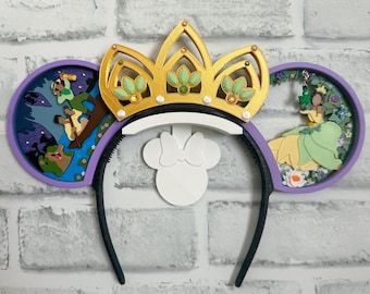 Könnte beinhalten: Lila und grüne Mäuseohren mit einer goldenen Krone und einem weißen Stirnband. Die Ohren zeigen eine Szene aus dem Film 'Die Prinzessin und der Frosch'.