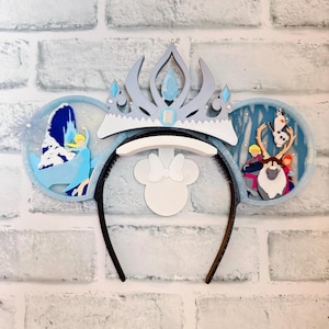 May include: A themed headband featuring a crown and two circular ear accents. The ears depict scenes from a popular animated film, with characters and icy landscapes in shades of blue, white, and gray. A white bow is centered.