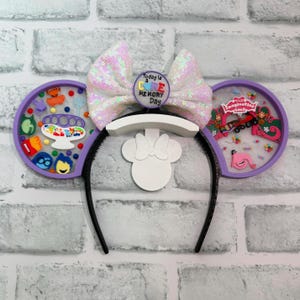 May include: A black headband with purple ears, a white bow, and a white Minnie Mouse silhouette. The ears have colourful designs and the bow is covered in iridescent sequins. The centre of the bow has a button that says "Today is a Core Memory Day."