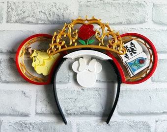 Könnte beinhalten: Rote und goldene Mickey Mouse Ohren mit einem Beauty and the Beast Thema. Die Ohren zeigen eine goldene Krone mit einer roten Rose, eine gelbe Belle Figur und ein Buch mit dem Text "Tale as old as time".