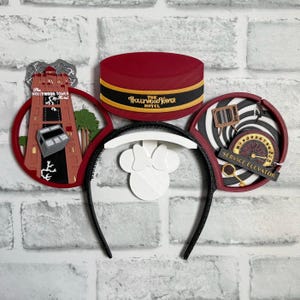 Op de afbeelding: Rode en zwarte Mickey Mouse oren met een Hollywood Tower Hotel ontwerp op het ene oor en een Service Elevator ontwerp op het andere oor. De oren zijn bevestigd aan een zwart hoofdband.