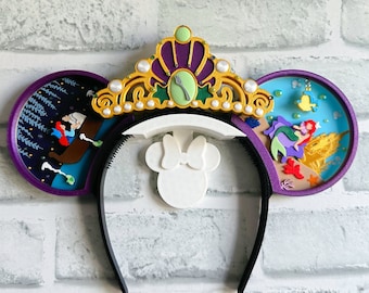 Könnte beinhalten: Lila und blaue Minnie Mouse Ohren mit einer goldenen Krone, die mit einer grünen Muschel und einer Gabel verziert ist. Die Ohren haben eine weiße Mitte mit einer Schleife und auf der einen Seite eine Szene mit Arielle und Ursula und auf der anderen Seite eine Szene mit der kleinen Meerjungfrau.
