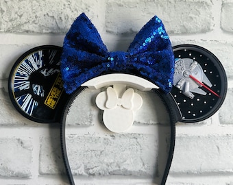 Puede incluir: Orejas de Minnie Mouse negras y blancas con temática de Star Wars con un lazo azul de lentejuelas. La oreja izquierda presenta una escena de Star Wars: El Imperio Contraataca con el texto "Nunca me digas las probabilidades". La oreja derecha presenta un modelo del Halcón Milenario.