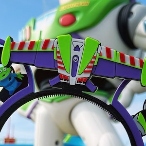 May include: Purple and green plastic headband with a 3D design of Buzz Lightyear's spaceship from the Toy Story movie. The headband features a green alien character on one side and a Buzz Lightyear character on the other side.