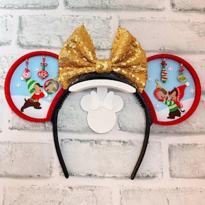 May include: A black headband with attached Minnie Mouse ears. The ears are decorated with a gold sequined bow and two circular ornaments with a red border. The ornaments feature a winter scene with cartoon characters and Christmas decorations.