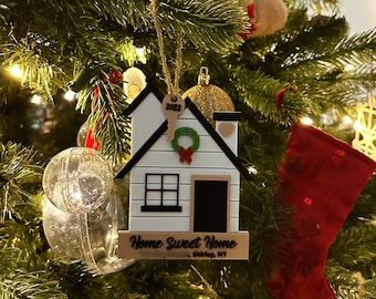 Puede incluir: Adorno navideño de madera blanca en forma de casa con una puerta y ventanas negras, una corona verde con un lazo rojo, una llave dorada con el año 2022 en ella y un corazón dorado. El adorno tiene un letrero de madera que dice "Home Sweet Home Shirley, NY".