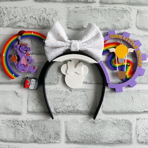 May include: Purple and white Mickey Mouse ears with a rainbow and a purple dragon on one side and a purple gear with a rainbow and a hot air balloon on the other side. The text "One Little Spark" is on the gear.