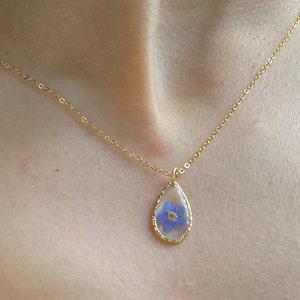 May include: A gold chain necklace with a teardrop-shaped pendant featuring a pressed blue flower.
