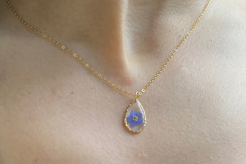 May include: A gold chain necklace with a teardrop-shaped pendant featuring a pressed blue flower.