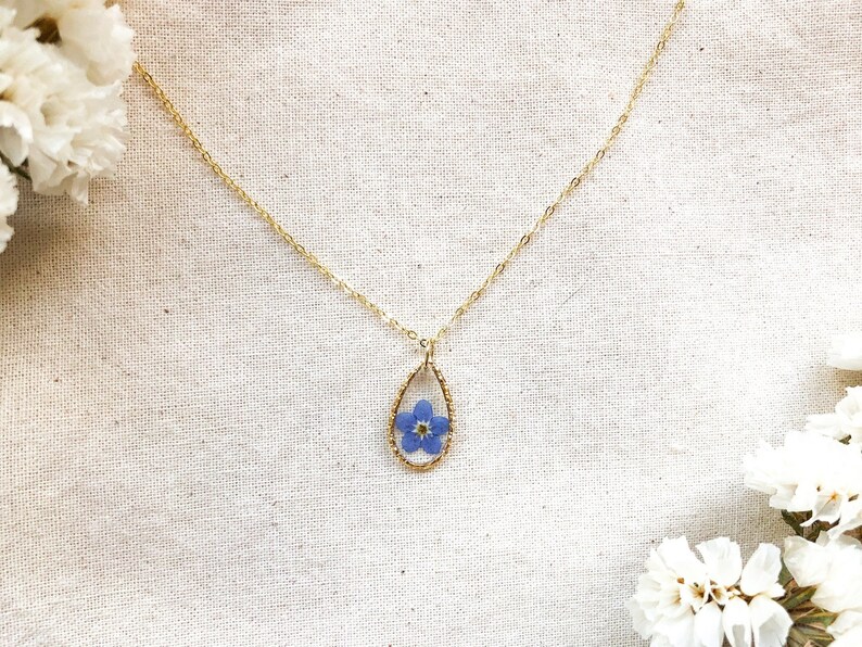 May include: A delicate gold chain necklace with a teardrop-shaped pendant featuring a pressed forget-me-not flower.