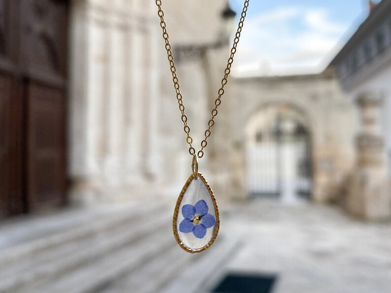 May include: A gold chain necklace with a teardrop-shaped pendant. The pendant is clear and contains a pressed blue flower.