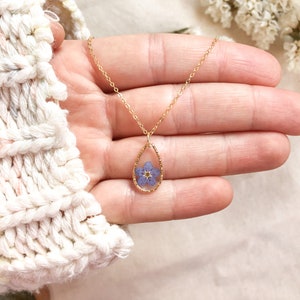 May include: A gold chain necklace with a teardrop-shaped pendant featuring a pressed purple flower inside.