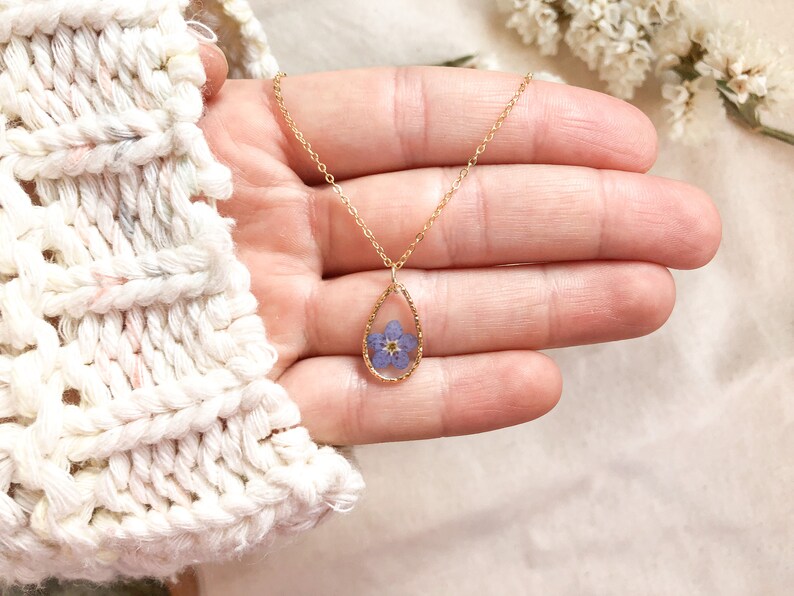 May include: A gold chain necklace with a teardrop-shaped pendant featuring a pressed purple flower inside.